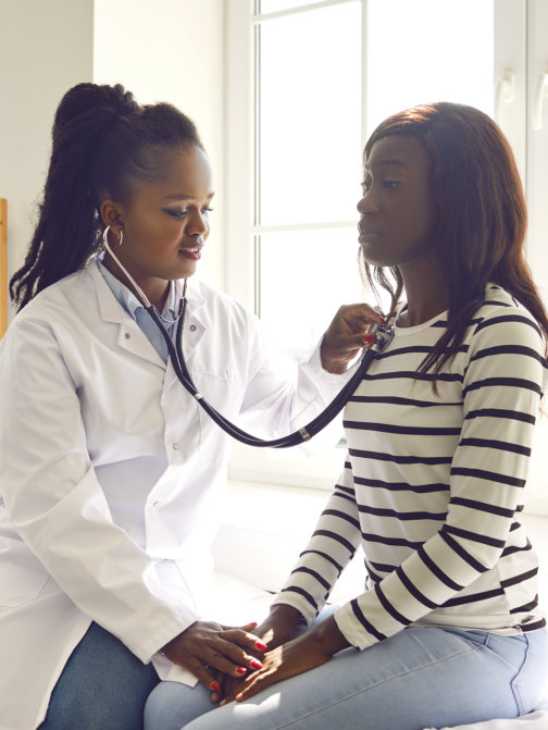 African american doctor with stethoscope examining young woman's lungs, breathing and heartbeat sitting on couch in exam room during medical checkup in clinic
